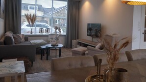 A modern living room with a sofa, a dining table with a tray, and a view of the outside through large windows. - Julia's Holidayhome (Egmond-Binnen)