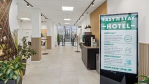 Una moderna zona de recepción de hotel con un cartel digital, mostrador de recepción y plantas en macetas.