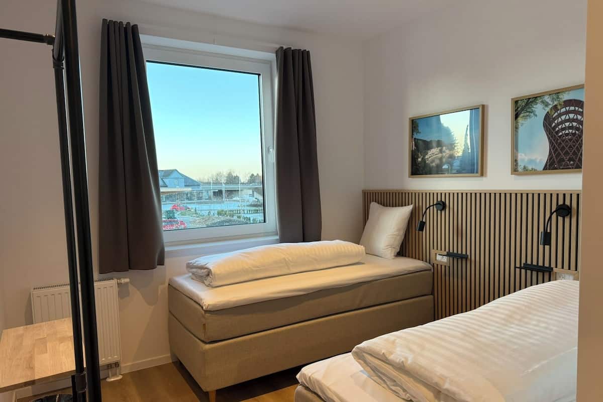 Ett hotellrum med en säng, ett fönster med gardiner, ett element, en papperskorg och två inramade tavlor på väggen.