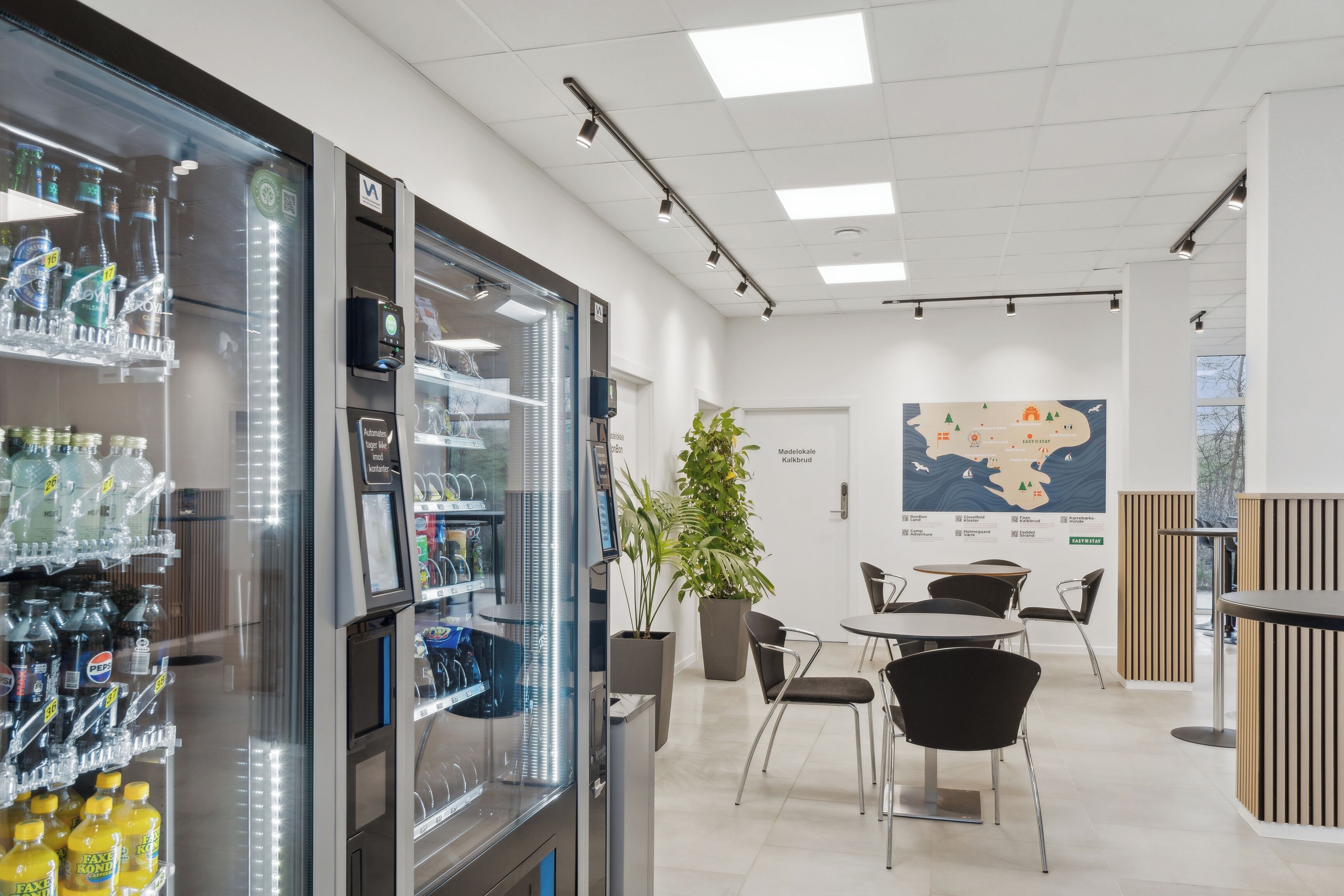 Un hall d’hôtel moderne doté d’un espace de service en libre-service pour les boissons, d’une grande carte affichée au mur et d’un coin salon avec des tables et des chaises.