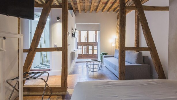 Una habitación con vigas de madera a la vista, una cama, un sofá, una pequeña mesa redonda y vistas al balcón a través de un ventanal.