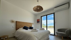 Un dormitorio moderno con una cama grande, un cabecero de madera, un taburete pequeño, un sillón gris y vistas al exterior a través de un ventanal.