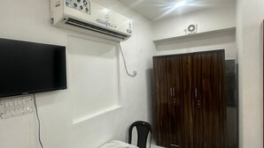 A small, well-lit room with a white air conditioner, a flat-screen TV mounted on the wall, a wooden wardrobe, a marble-topped table, and two plastic chairs. - Hotel Star Marigold (New Delhi)