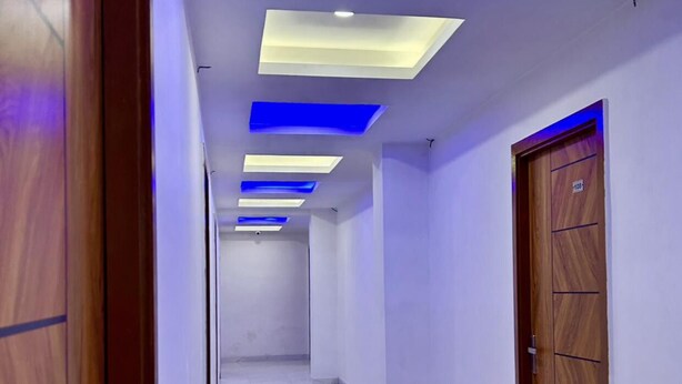 A hallway with a wooden door on the left, a blue-lit ceiling, and a white tiled floor. - Hotel Star Marigold (New Delhi)
