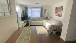 Un vaste espace de vie comprenant une longue table en bois, un canapé, une petite table basse et un tapis. On y trouve une fenêtre avec des rideaux, une plante et un tableau accroché au mur.