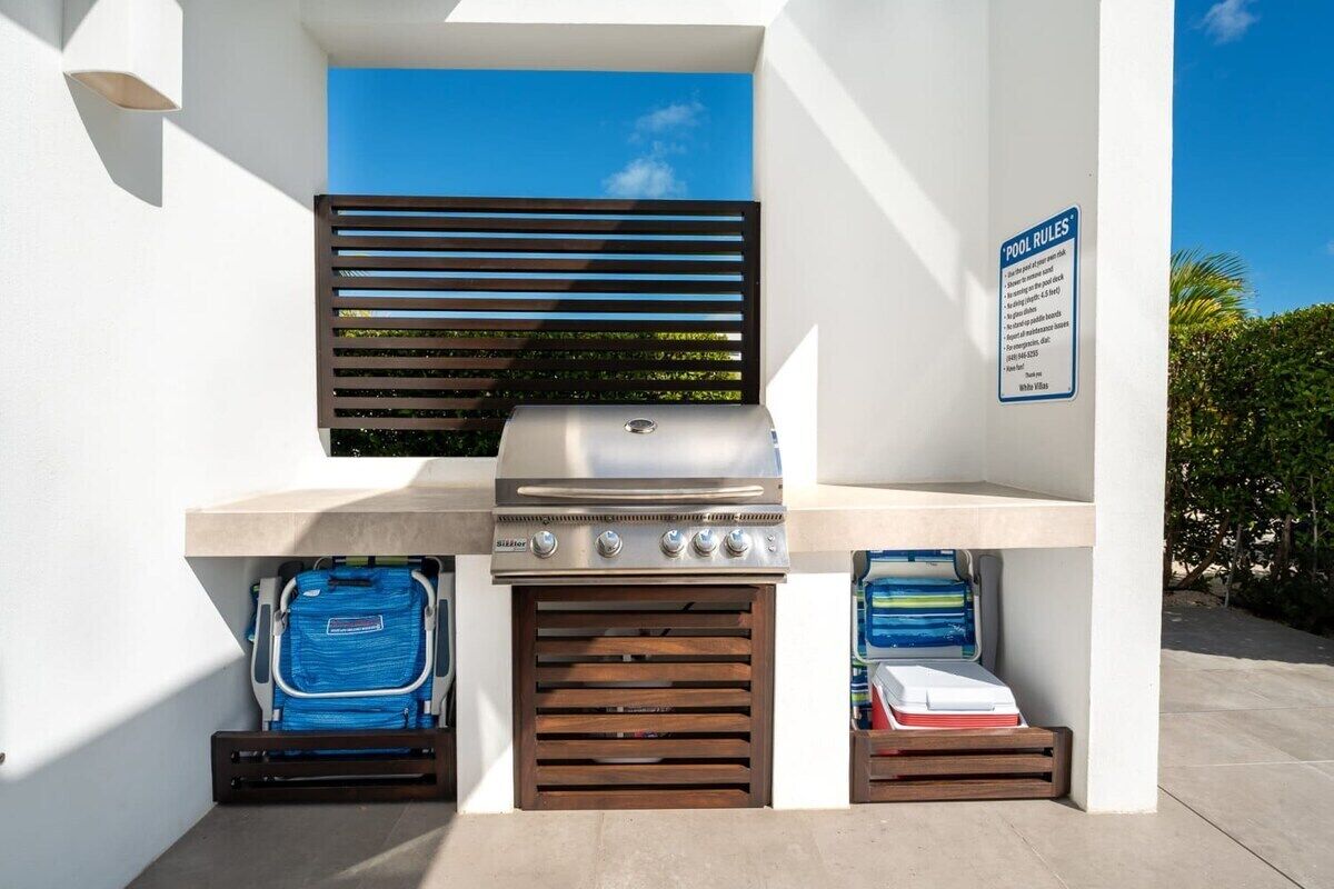 An outdoor grill area with a blue cooler and a white cooler, a 'POOL RULES' sign, and a clear blue sky in the background.