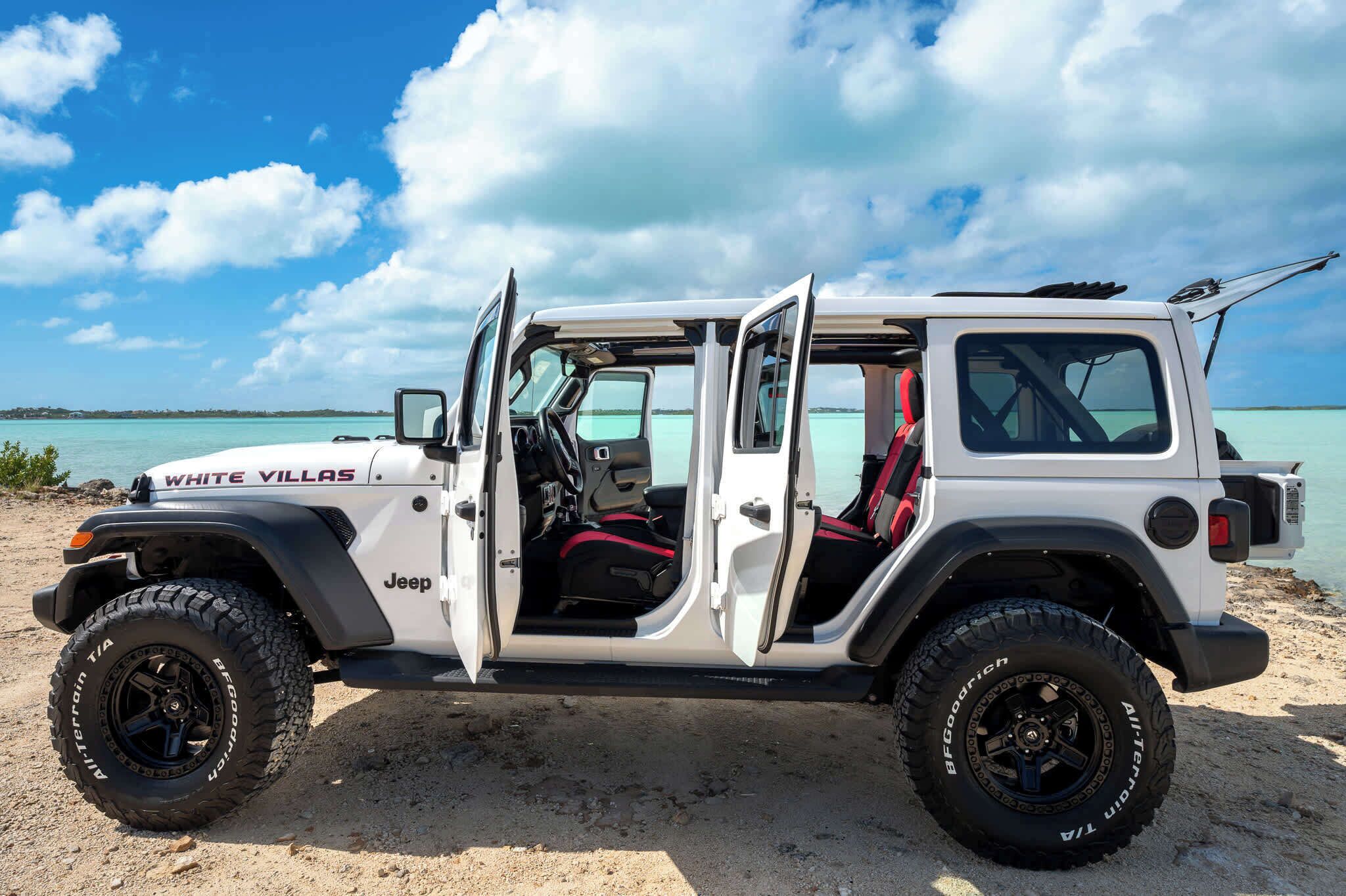 Un Jeep blanco con detalles interiores rojos estacionado en una playa arenosa, con aguas turquesas al fondo.