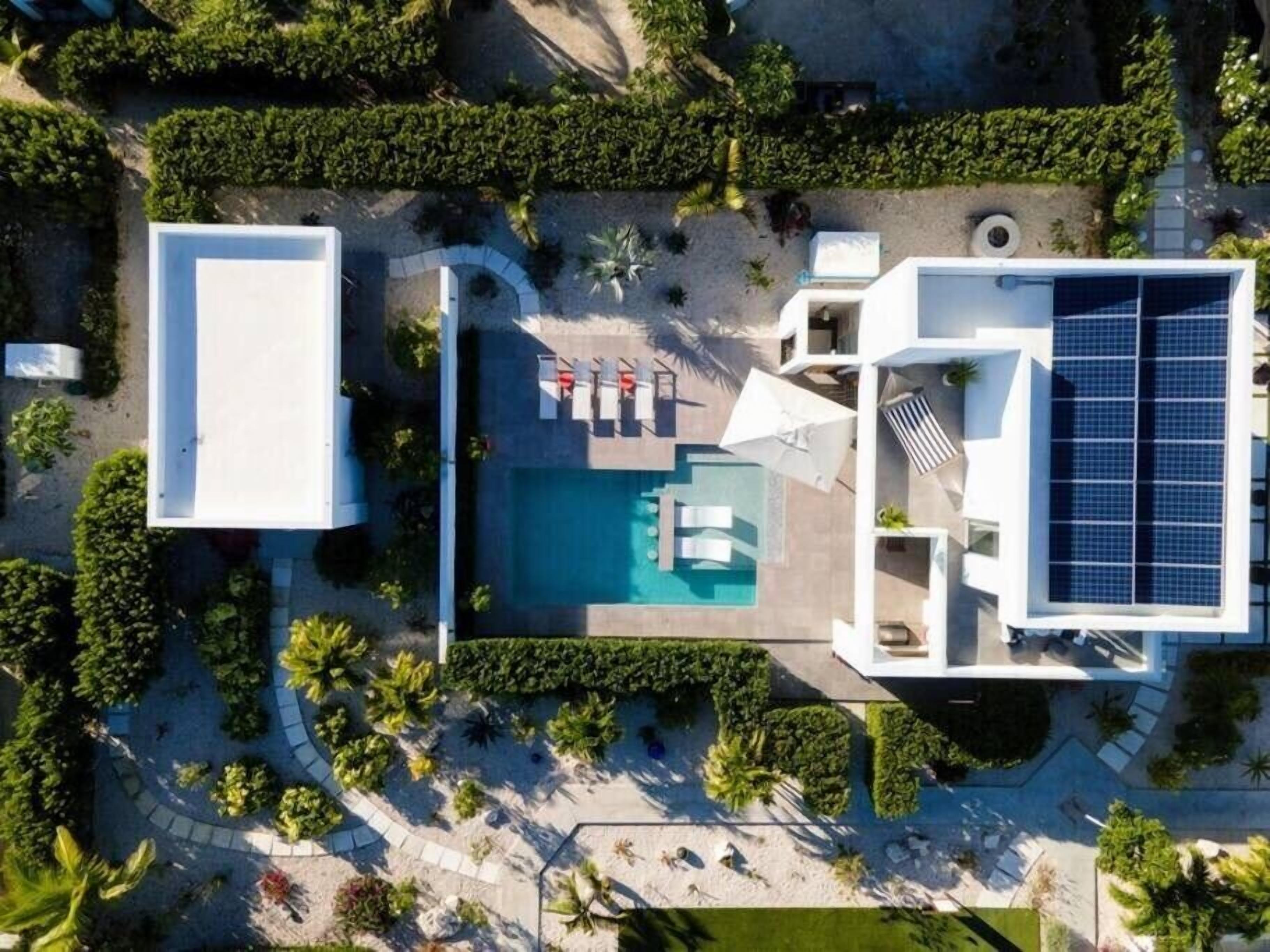 An aerial view of a modern resort with a central swimming pool, surrounded by sun loungers and umbrellas, and solar panels on the right.