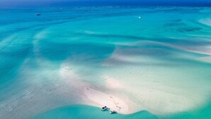 Ein Luftbild zeigt einen Strand mit klarem türkisblauem Wasser, eine kleine Sandinsel mit einigen Booten und ein Schiff in der Ferne.