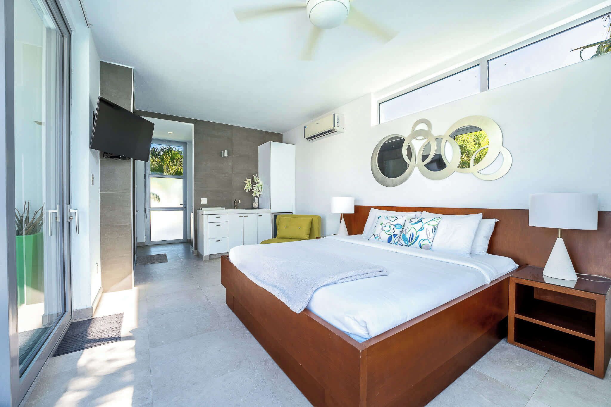 Une chambre moderne avec un grand lit, une table de chevet, un téléviseur fixé au mur et une vue sur la cuisine et le balcon.