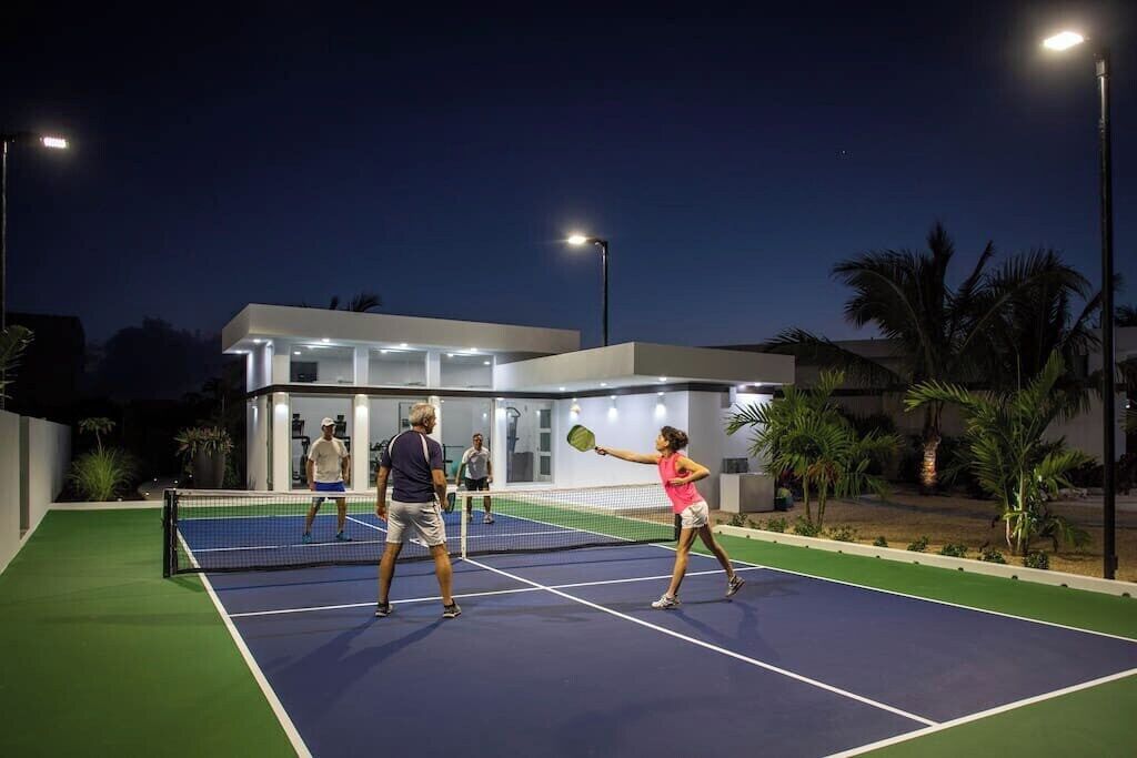 Una cancha de tenis al aire libre con una red, rodeada por un edificio moderno con ventanas de vidrio y techo plano.
