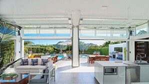 A modern kitchen with a large island, a dining area with outdoor seating, and a view of a pool and mountains. - Family Beachside Villa With Hotel Amenities v7 (Providenciales)