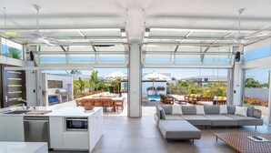 A modern open-plan living space with a kitchen, dining area, and seating area, featuring large windows and a view of the outdoors. - New Seaside Vacation Villa With Hotel Amenities v9 (Providenciales)