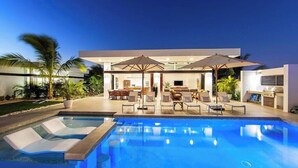 A modern outdoor pool area with lounge chairs and umbrellas, a dining area with tables and chairs, and a well-maintained garden with palm trees. - New Seaside Vacation Villa With Hotel Amenities v9 (Providenciales)