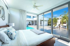 Camera d'albergo moderna con un letto grande, un ventilatore a soffitto e vista su piscina e area relax all'aperto.