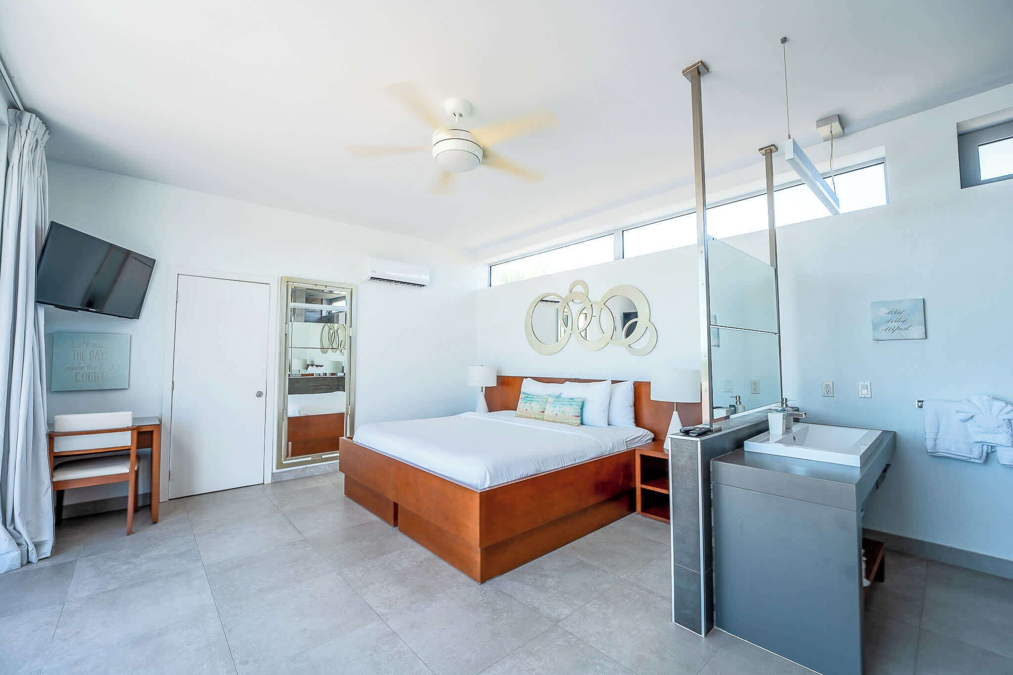 Une chambre moderne avec un grand lit, une coiffeuse intégrée, un ventilateur de plafond et un téléviseur fixé au mur.