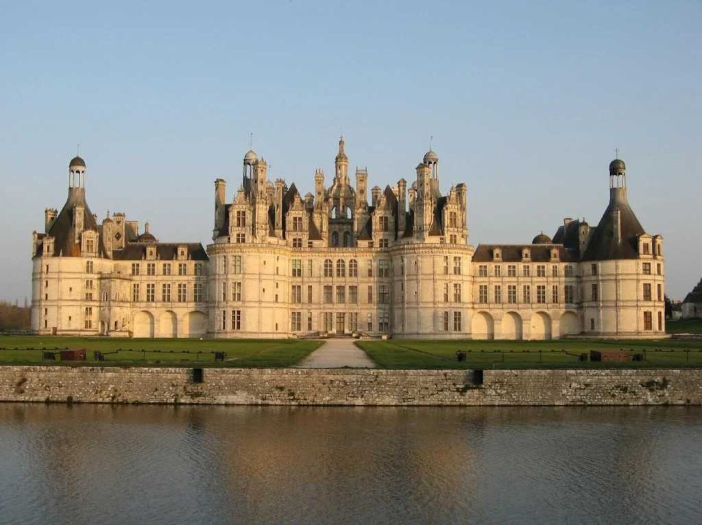 A large, historic building with multiple towers and a symmetrical facade, surrounded by a moat and green lawns.