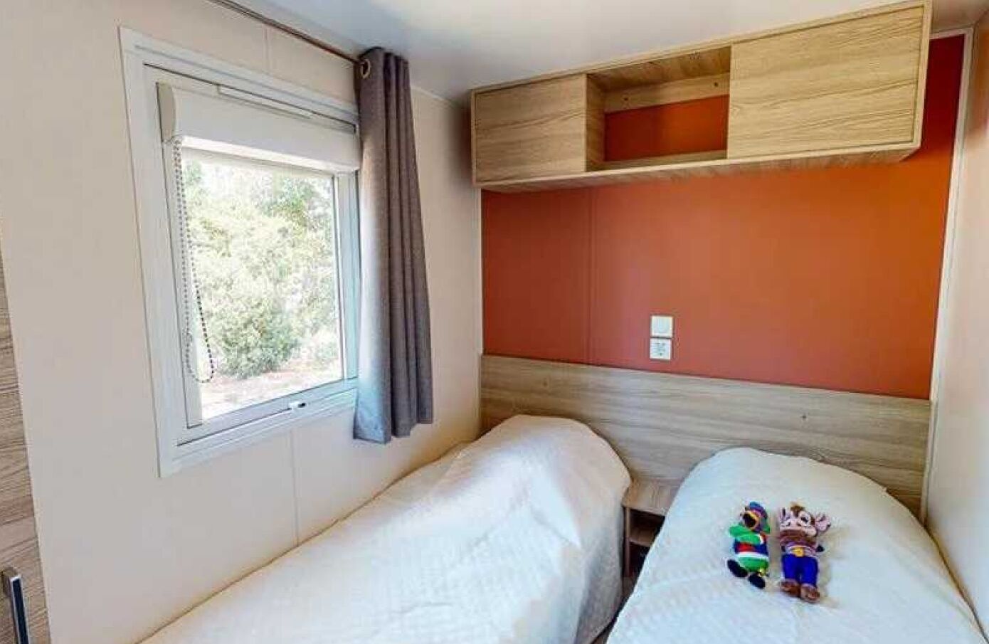 Une chambre avec deux lits, une fenêtre avec des rideaux, une tête de lit en bois et une étagère au-dessus des lits.