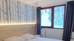 A bedroom with a large bed, a wall-mounted light panel, a window with curtains, and a view of snow-covered trees. - Bright and spacious apartment for 6 people - View of the Flaine Forum (Arâches-la-Frasse)