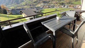 A balcony with wicker furniture and a table, overlooking a green valley with houses and hills. - Apartment Mountain Bliss (Kaprun)