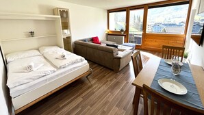 A room with a bed, a sofa, a dining table with a plate and glass, and a window with a view of buildings. - Apartment Mountain Bliss (Kaprun)