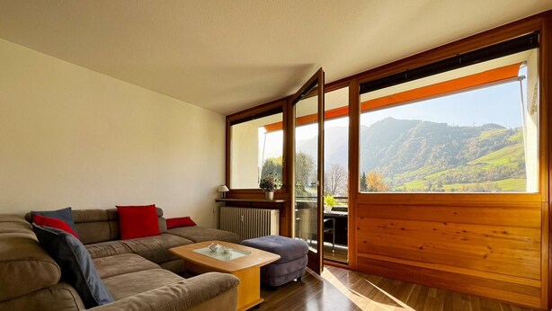 A living room with a sofa, a coffee table, and a view of a mountainous landscape through large windows. - Apartment Mountain Bliss (Kaprun)