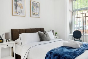 A bedroom with a bed, white nightstands, a lamp, a window with a view, and two framed artworks on the wall.