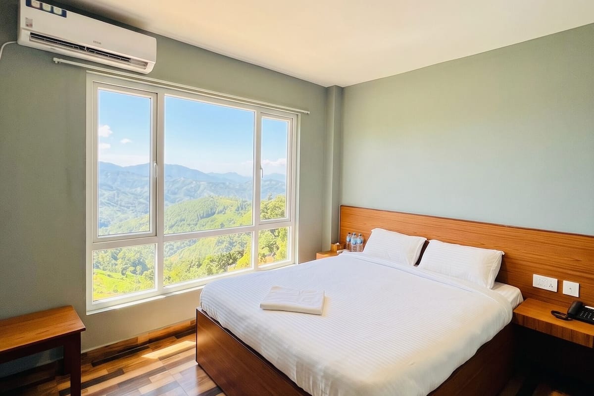 Habitación de hotel con una cama grande, suelo de madera, una mesita de noche y una ventana con vistas a la montaña.