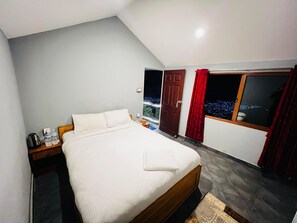 A neatly made bed in a room with a view of a cityscape at night, a door, and a window with red curtains. - Tourist Land Resort (Dharan)