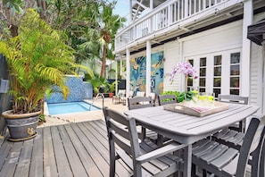 Une terrasse en bois avec une table et des chaises, une plante en pot, une piscine et un bâtiment blanc doté d’un balcon.