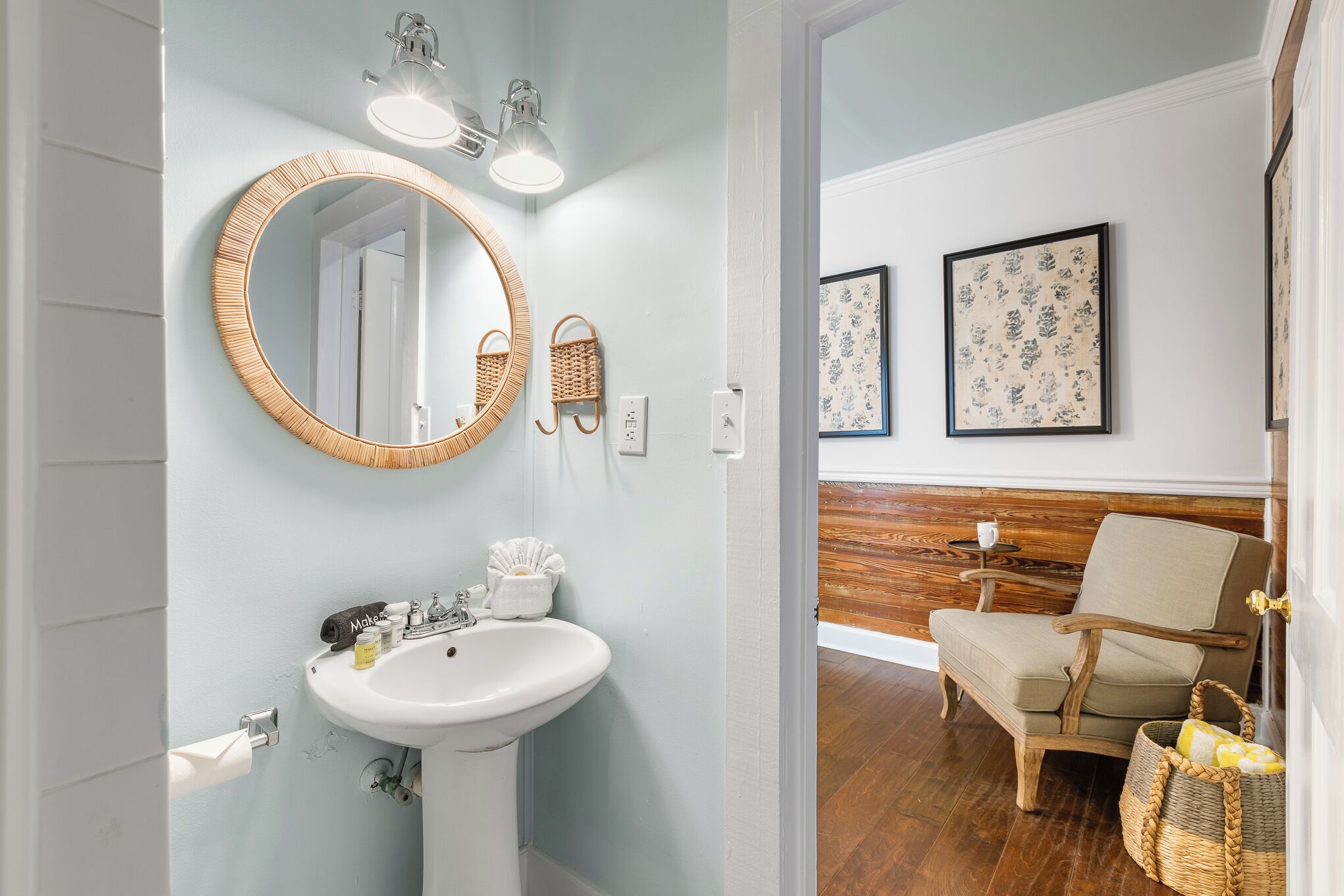 Un baño con un espejo redondo, lavamanos y luces empotradas en la pared. Hay una silla y una cesta en la habitación contigua.
