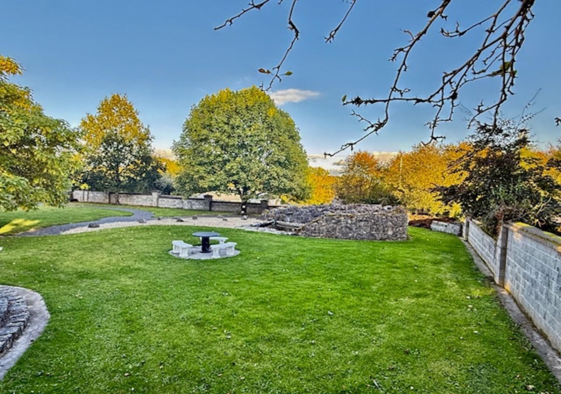 A well-maintained lawn with a circular seating area, stone walls, and trees with autumn foliage.