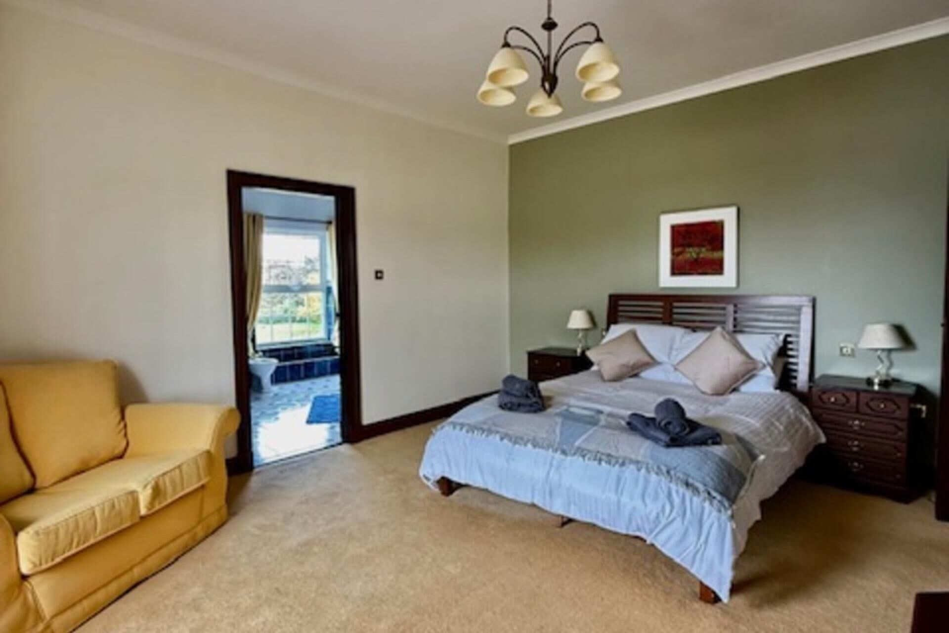 Un dormitorio con una cama, un sillón amarillo, una mesita de noche, una lámpara y un cuadro en la pared.
