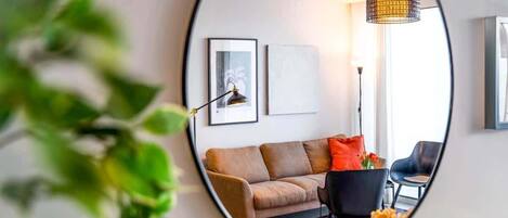 A round mirror reflecting a living room with a sofa, a chair, a lamp, and framed artwork on the wall.