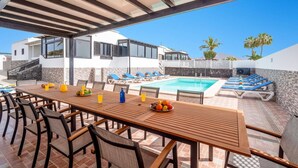 Uma área à beira da piscina com mesa e cadeiras de madeira, piscina e espreguiçadeiras.