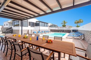 A poolside area with a wooden table and chairs, a swimming pool, and lounge chairs.