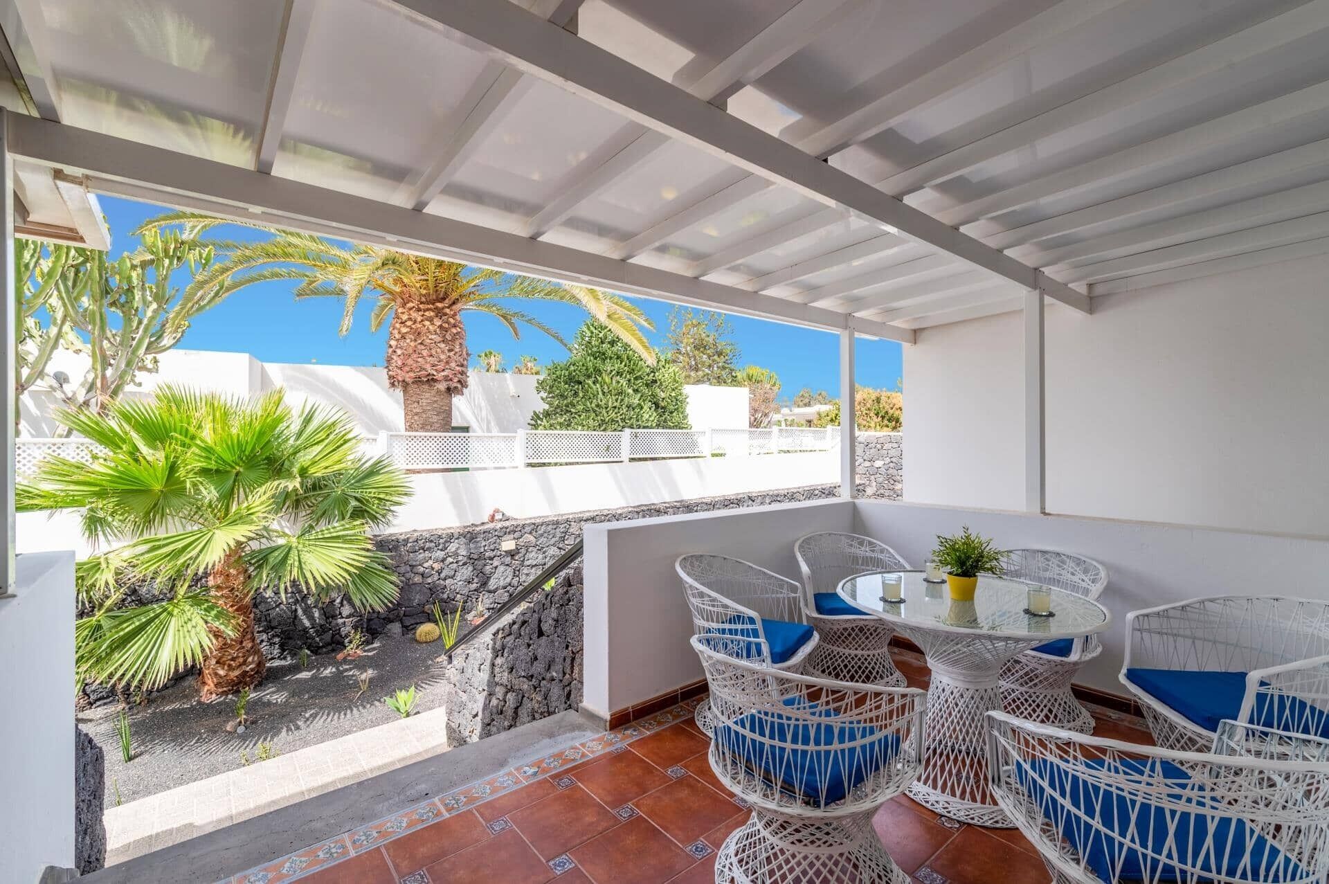 Un patio techado al aire libre con muebles de ratán blanco, una mesa redonda y vistas a un edificio blanco y palmeras.