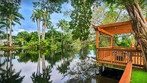A wooden deck over water, surrounded by lush greenery and palm trees. - Lakefront Lux Home • Pool_BBQ • Game Room & Kayak (Miami Springs)
