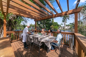 Una terraza de madera con zona de comedor cubierta, rodeada de vegetación y una masa de agua.