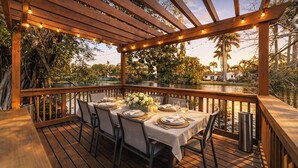 An outdoor dining area with a wooden deck, string lights, and a view of a lake and palm trees. - Lakefront Lux Home • Pool_BBQ • Game Room & Kayak (Miami Springs)