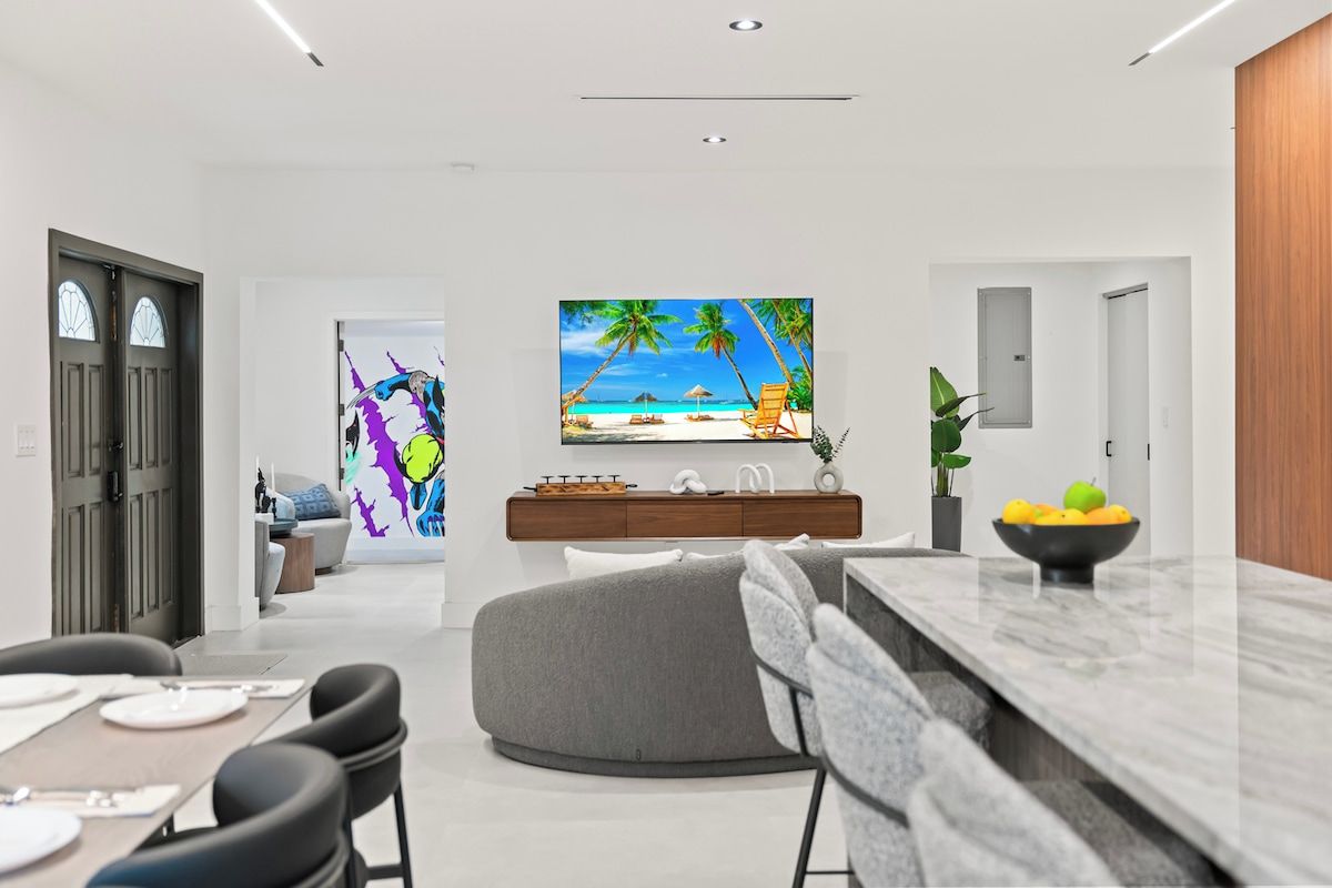 Una sala de estar moderna con vista al mar en la televisión, un comedor con mesa de mármol y un sofá gris.