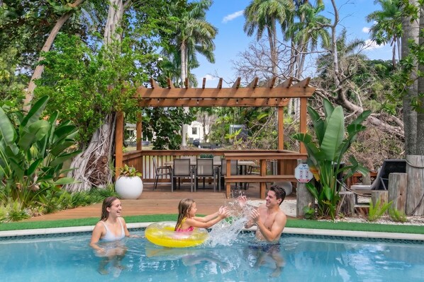 Piscina com pergolado de madeira, rodeada por vegetação tropical.