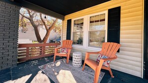 A porch with two wooden chairs and a small table, a brick wall, and a window with blinds.