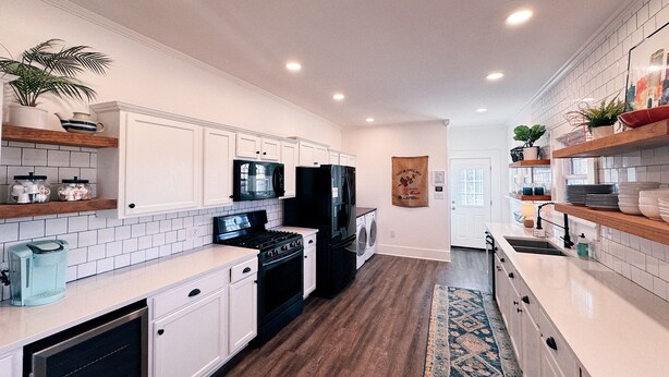 A modern kitchen with white cabinetry, a black refrigerator, a microwave, and a stove. There are wooden shelves with plants and kitchenware.
