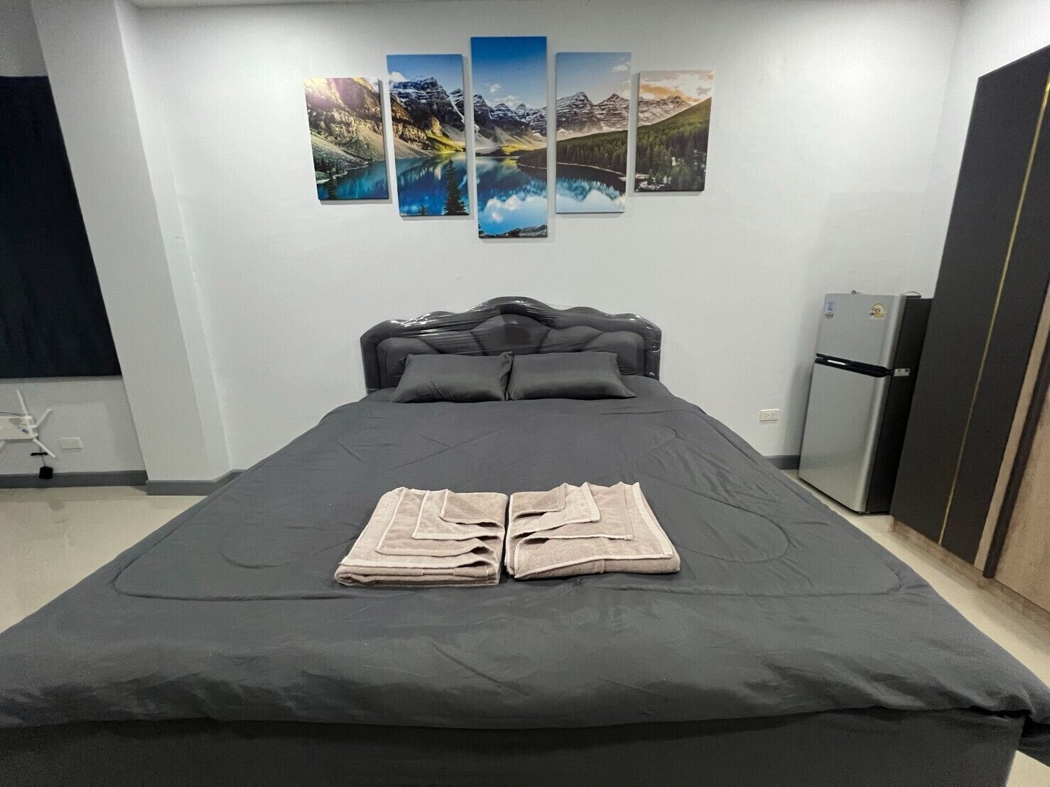 Un dormitorio con cama, televisor montado en la pared, refrigerador y un conjunto de pinturas.