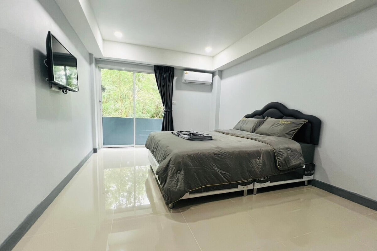A modern bedroom with a bed, a flat-screen TV mounted on the wall, and a large window with curtains.