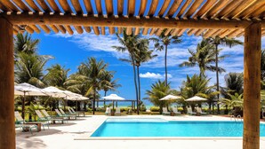 A pool area with lounge chairs and umbrellas, surrounded by palm trees and a clear blue sky. - Aldeia Beijupirá Pousada (Porto de Pedras)