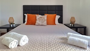 A neatly made bed with a textured white duvet, two orange pillows, and a black headboard. There are two bedside tables with decorative lamps and potted plants. - PRAIA DA ROCHA – ATLÂNTICO III (Portimão)
