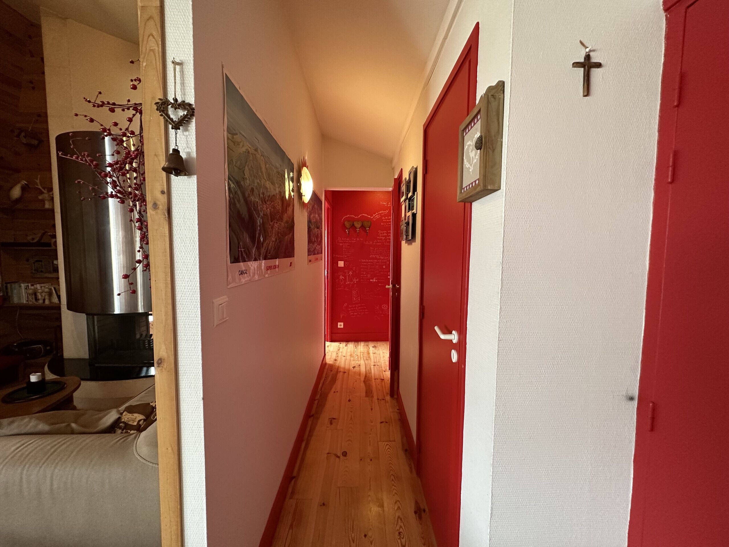 Un couloir étroit au plancher de bois, aux murs rouges et à la porte rouge au fond. Des tableaux encadrés et une cloche ornent le mur.