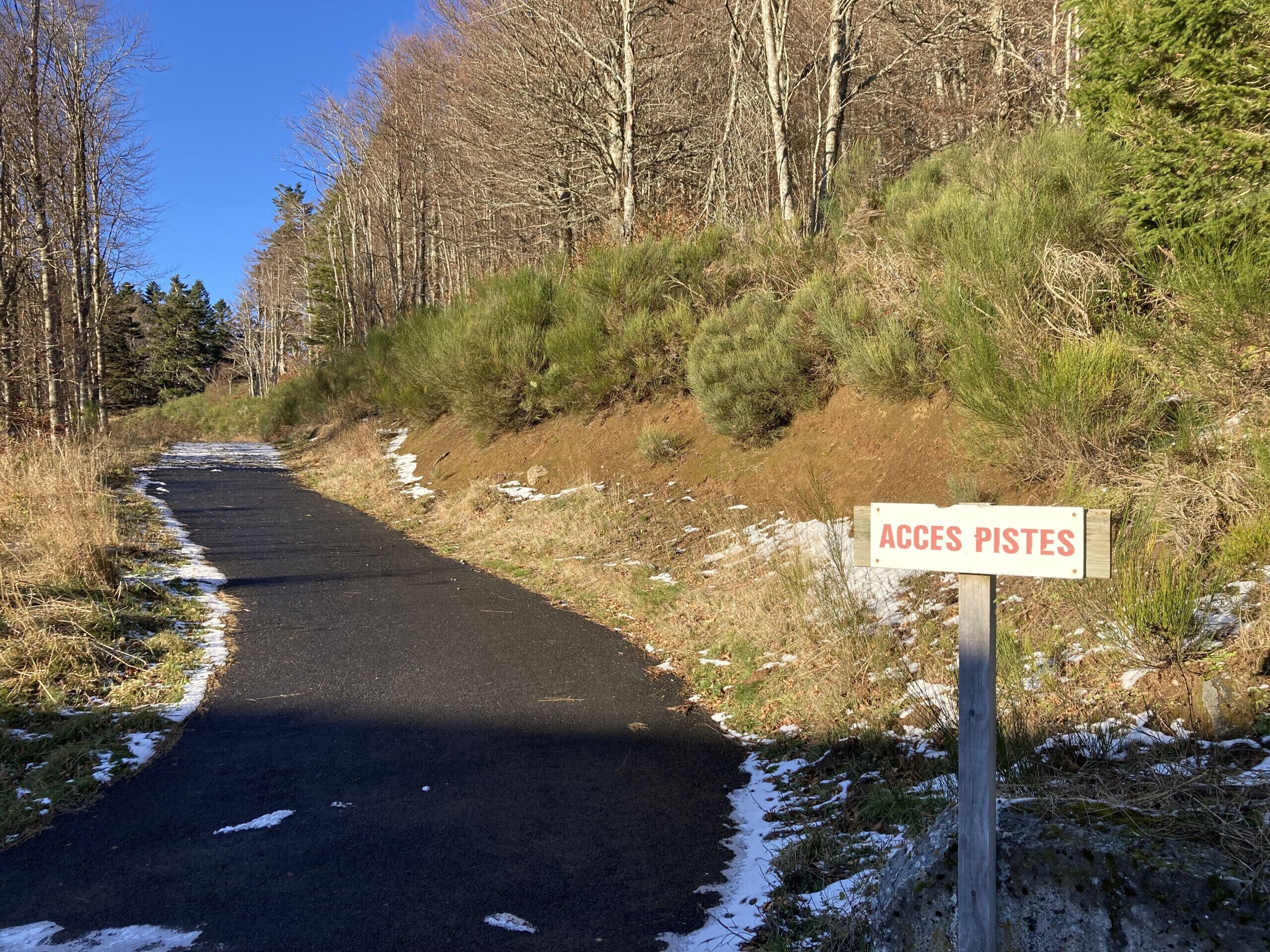 A paved path with a sign reading "ACCES PISTES" in a forested area.